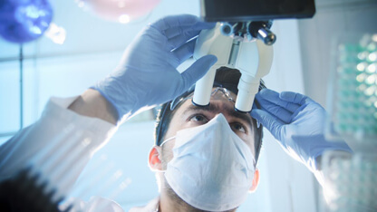 Scientist wearing a face mask and gloves examining samples through a microscope in a laboratory, with green-tinted lab equipment in the foreground.