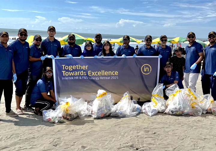 A group of 15 Intertek colleagues wearing dark blue tops and Intertek hats stand on a beach with the rubbish they have collected, with a sign that says "Together Towards Excellence". 