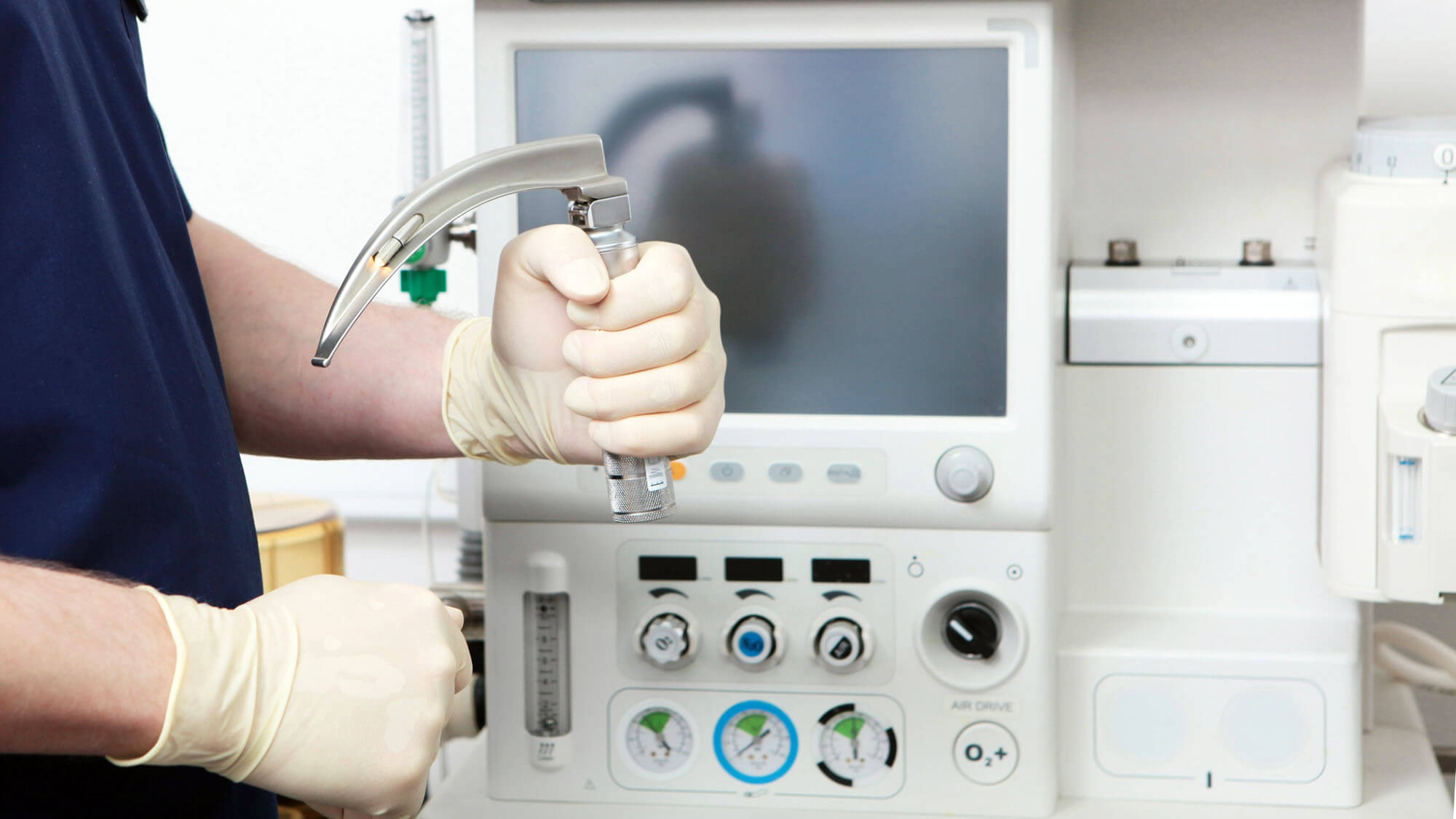 Doctor's hand in sterile gloves holds laryngoscope against background of modern anesthetic device in operating room